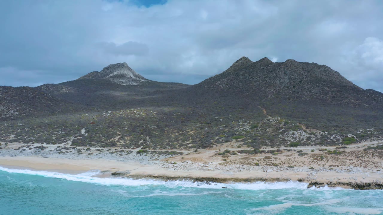 Aerial Los Cabos coastline with bright turquoise water in the foreground, a band of breaking surf, sandy beach, scrubby terrain, dark rocky hills, and no visible people.