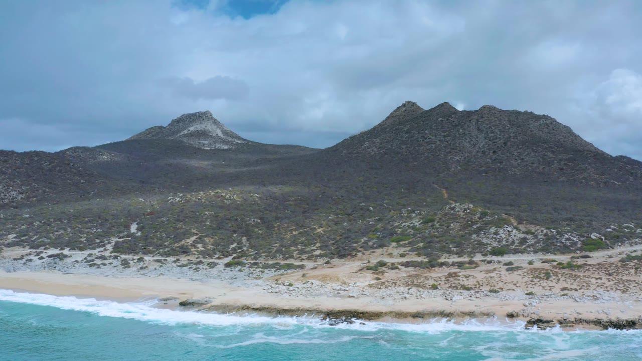 Los Cabos coastal landscape with surf washing across the lower frame, a light sand strip, scattered rocks and shrubs, broad mountain slopes, muted cloud light, and no visible people.