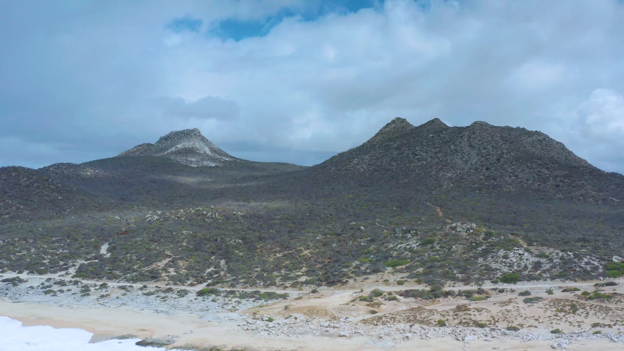 Los Cabos beach sand runs along the lower edge with low scrubland leading to twin rocky peaks under scattered clouds, with no visible people.
