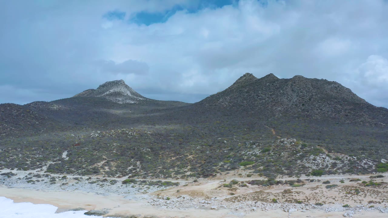 Los Cabos coastal scrub fills the midground while twin mountain peaks sit high on the horizon above pale sand, with no visible people.