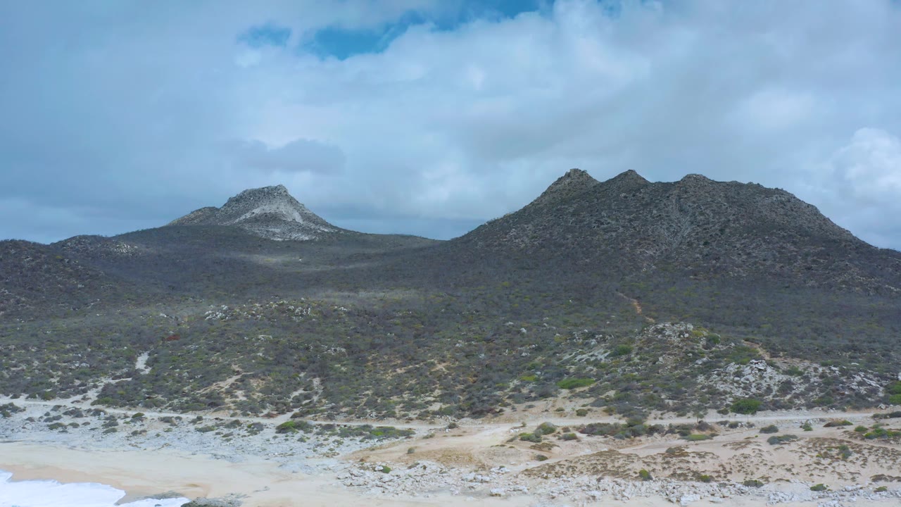 Los Cabos terrain shows a narrow sand strip below scrub-covered slopes and a dark twin-peaked ridge under bright cloud cover, with no visible people.