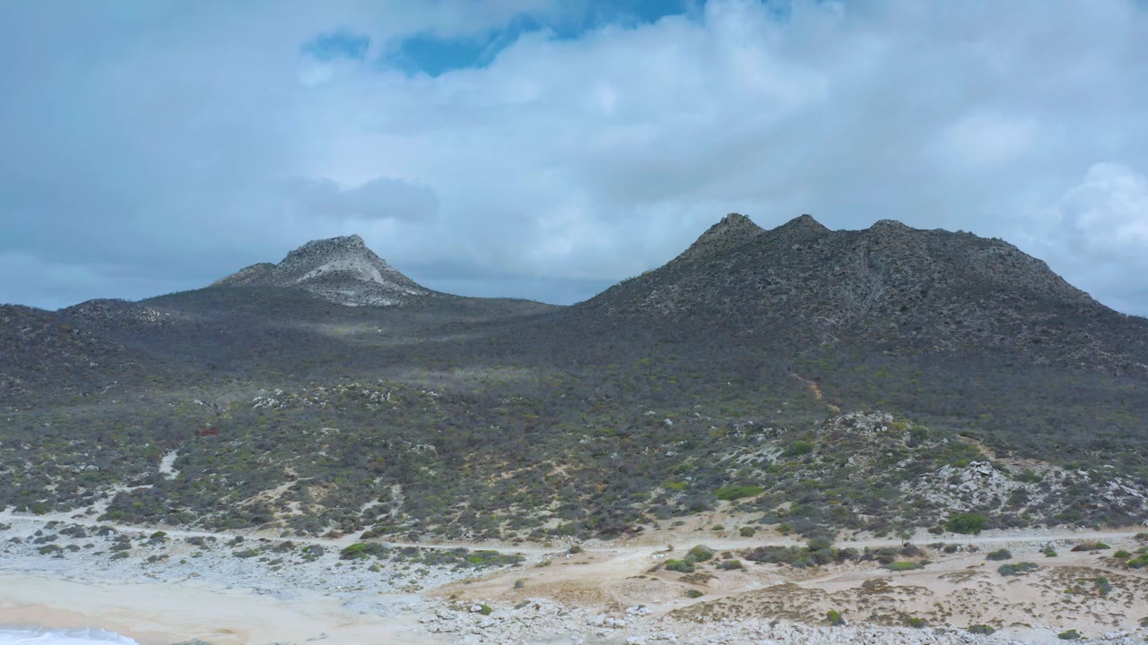 Los Cabos beach sand and light dune texture sit below a broad scrubland band and two rocky peaks beneath blue sky, with no visible people.