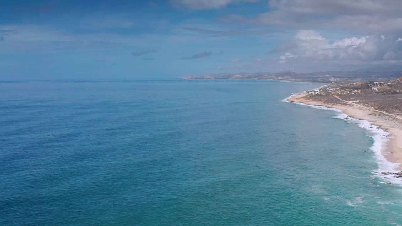 Aerial Los Cabos coastline frame with deeper blue water at left, turquoise water near shore, a pale beach curving along the right side, cloud cover near the horizon, and no visible people.