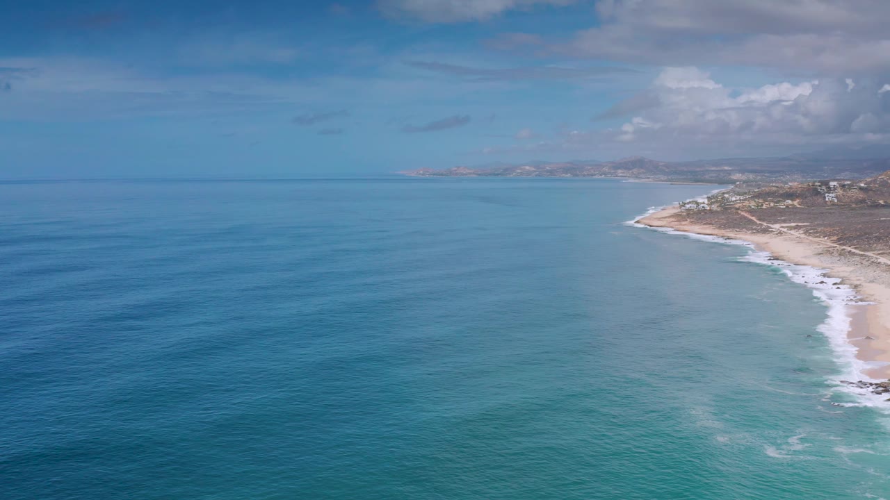 Los Cabos aerial coast view with smooth turquoise water in the midground, a curving sandy beach and foamy shorebreak on the right, low land in the distance, and no visible people.