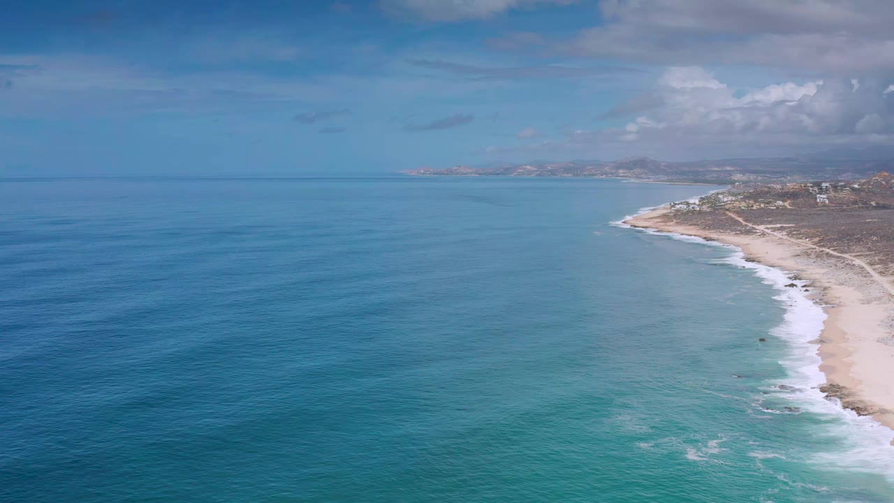Aerial Los Cabos beach view with rippled blue water in the foreground, white waves reaching the sand, distant coast on the horizon, and no visible people.