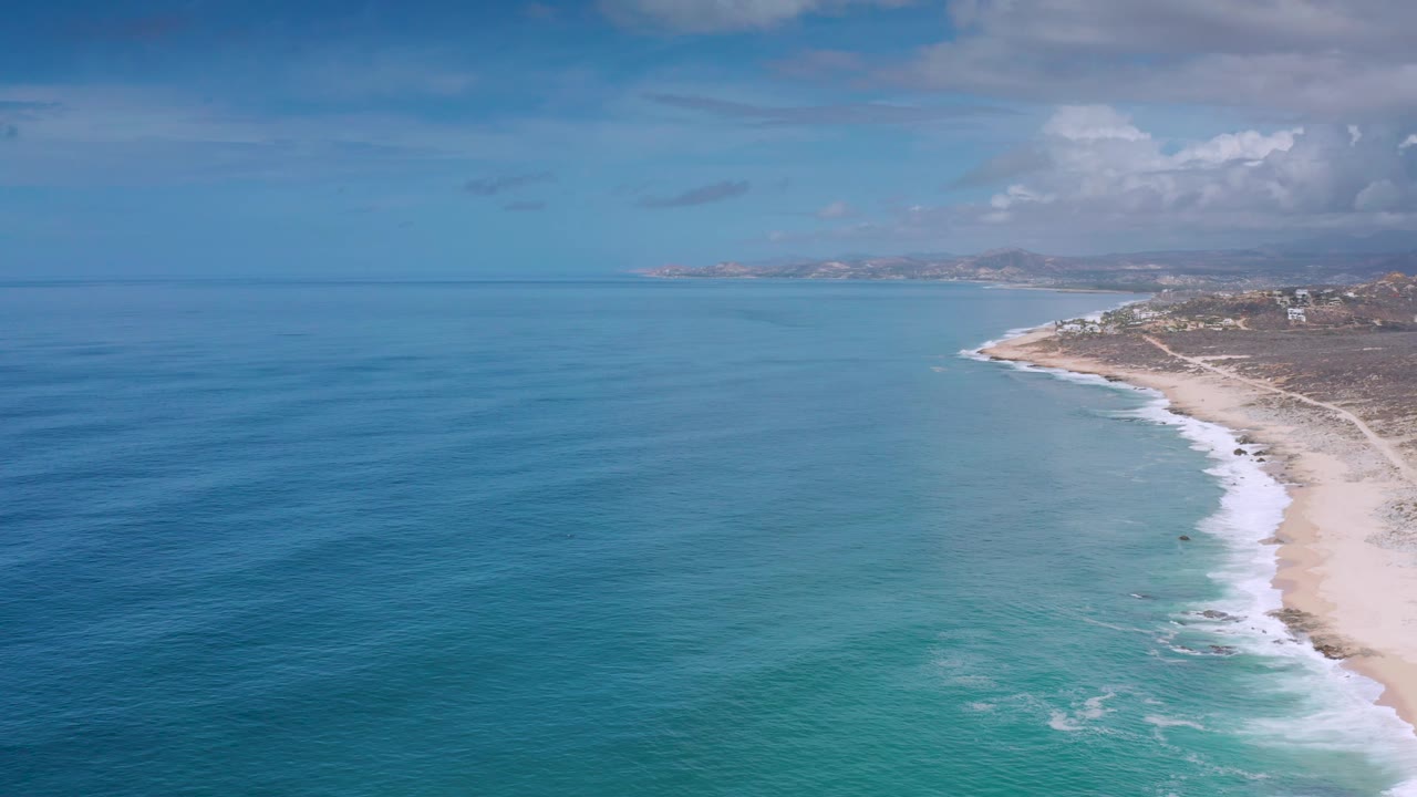 Wide aerial frame of Los Cabos coastline where turquoise shallows fade into deeper blue water, with the sandy beach and dry land running along the right edge and no visible people.