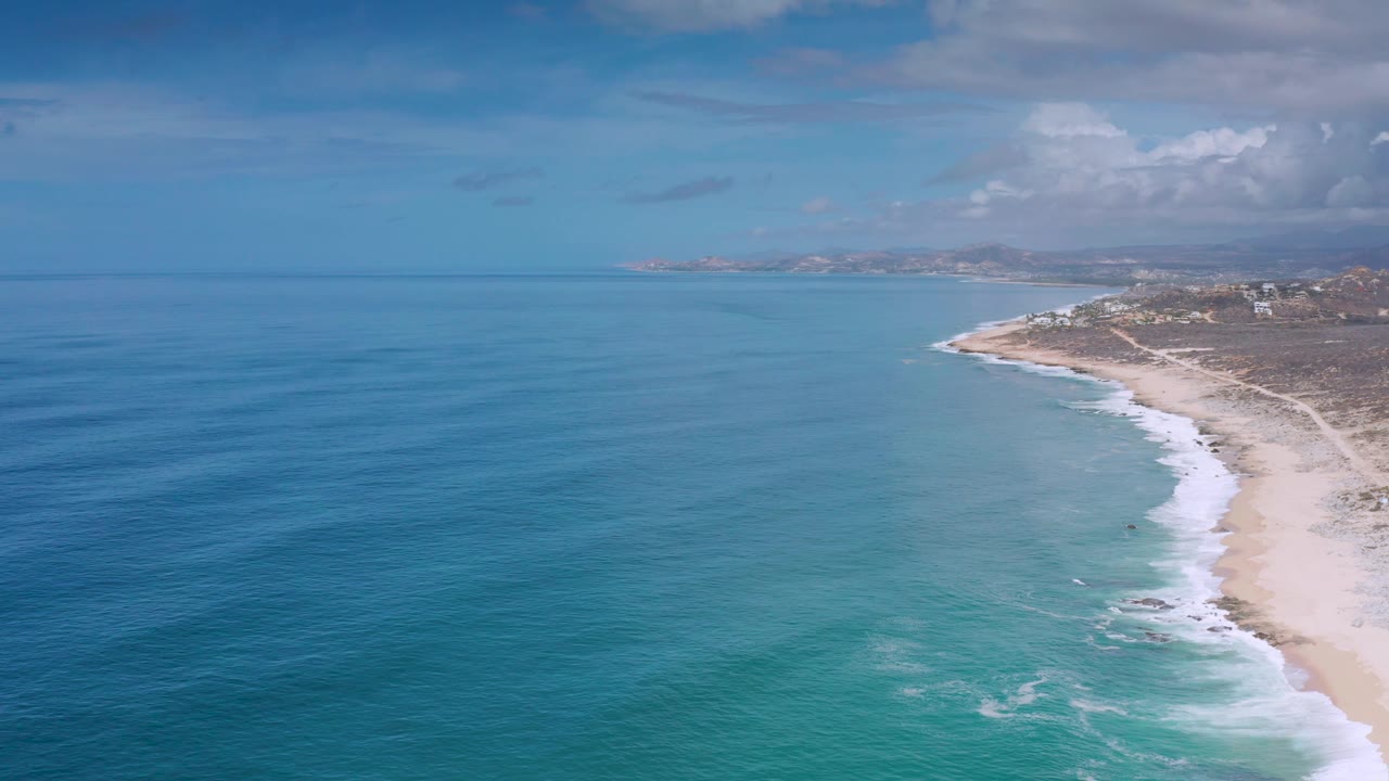 Aerial view of an empty Los Cabos shoreline with calm blue ocean to the left, pale sand to the right, dry inland terrain, and bright clouds above.