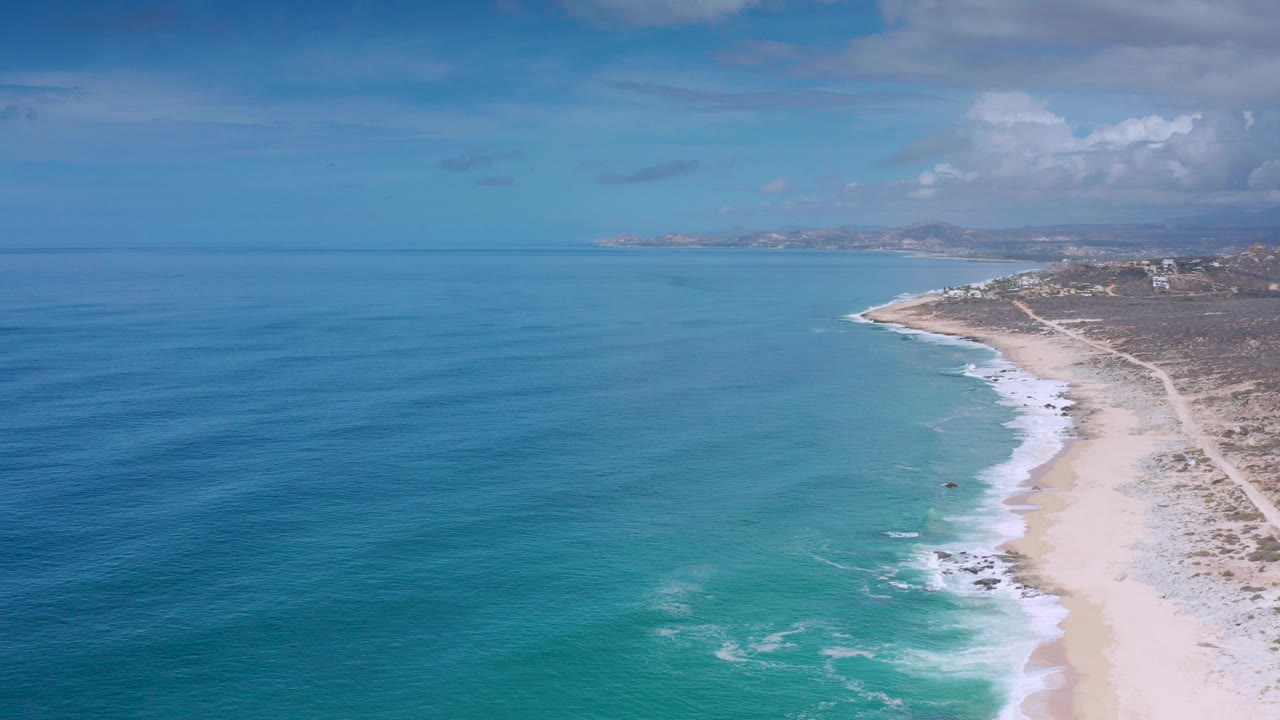 Opening aerial frame over the Los Cabos coast, with blue ocean on the left, surf reaching a pale sand beach, scrub inland, distant land on the horizon, and no visible people.