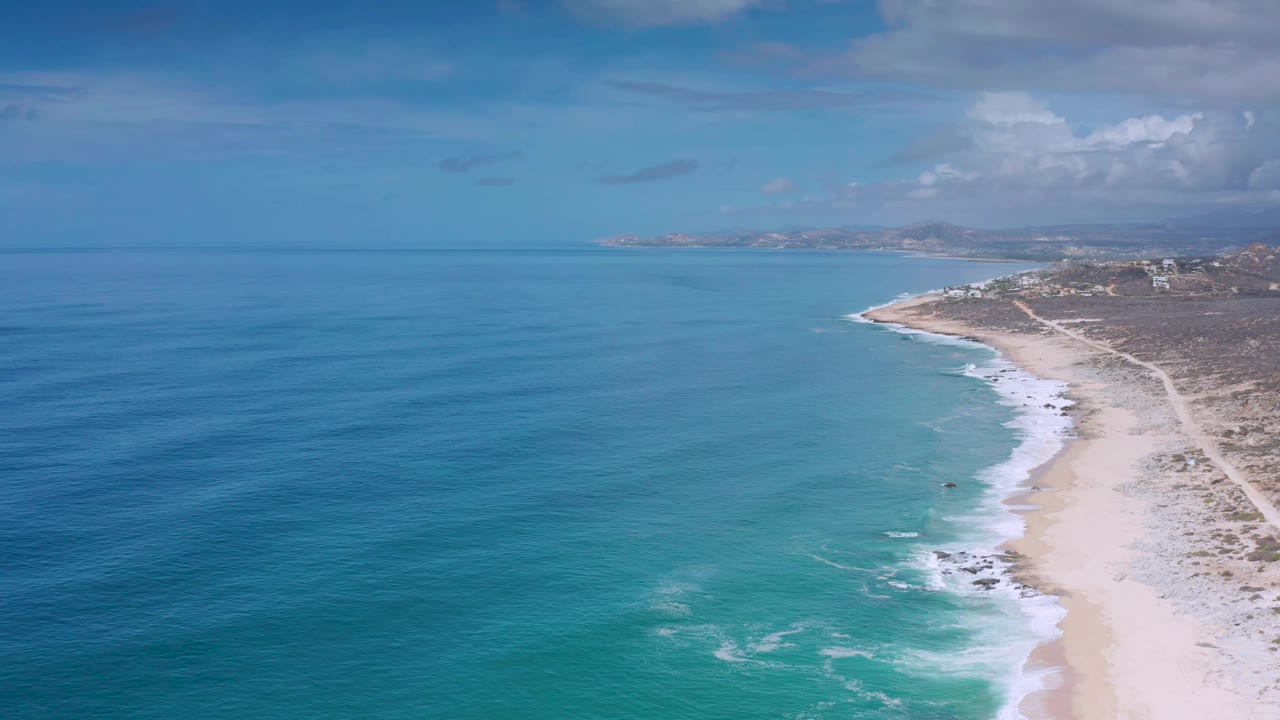 Aerial Los Cabos shoreline view with more open water in the foreground, white wave foam marking the beach edge, a dirt track inland, and no visible people.