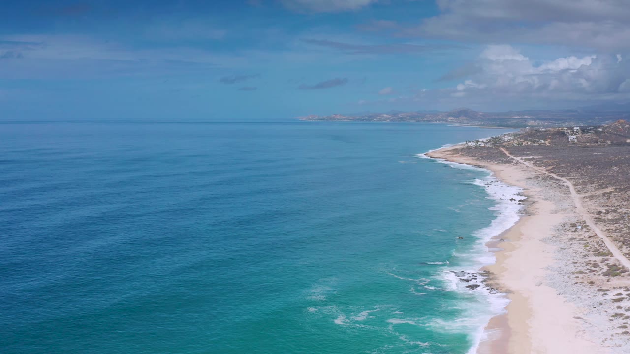 High oblique aerial frame of the Los Cabos coast, with turquoise shallows spreading toward the beach, small white breakers, dry coastal scrub to the right, and no visible people.