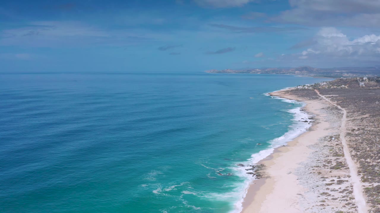 Drone view in Los Cabos, Mexico, with rippled blue water, turquoise surf, scattered dark rocks along the sand, coastal scrub, and no visible people.