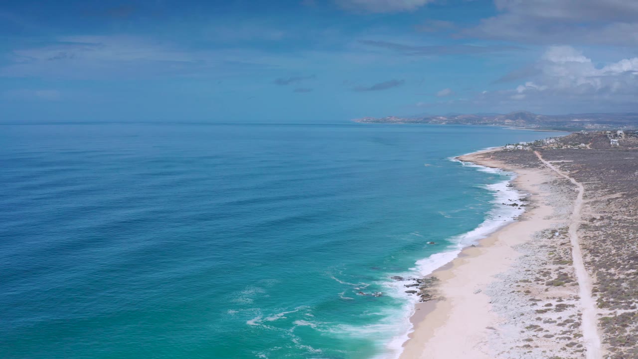 High aerial frame in Los Cabos, Mexico, showing long blue ocean ripples, a curving sandy shoreline, surf bands, dry coastal vegetation, and no visible people.