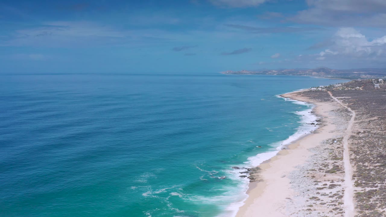 Aerial beach view in Los Cabos, Mexico, with calm blue ocean filling most of the frame, surf along the sand, scrubland to the right, and no visible people.