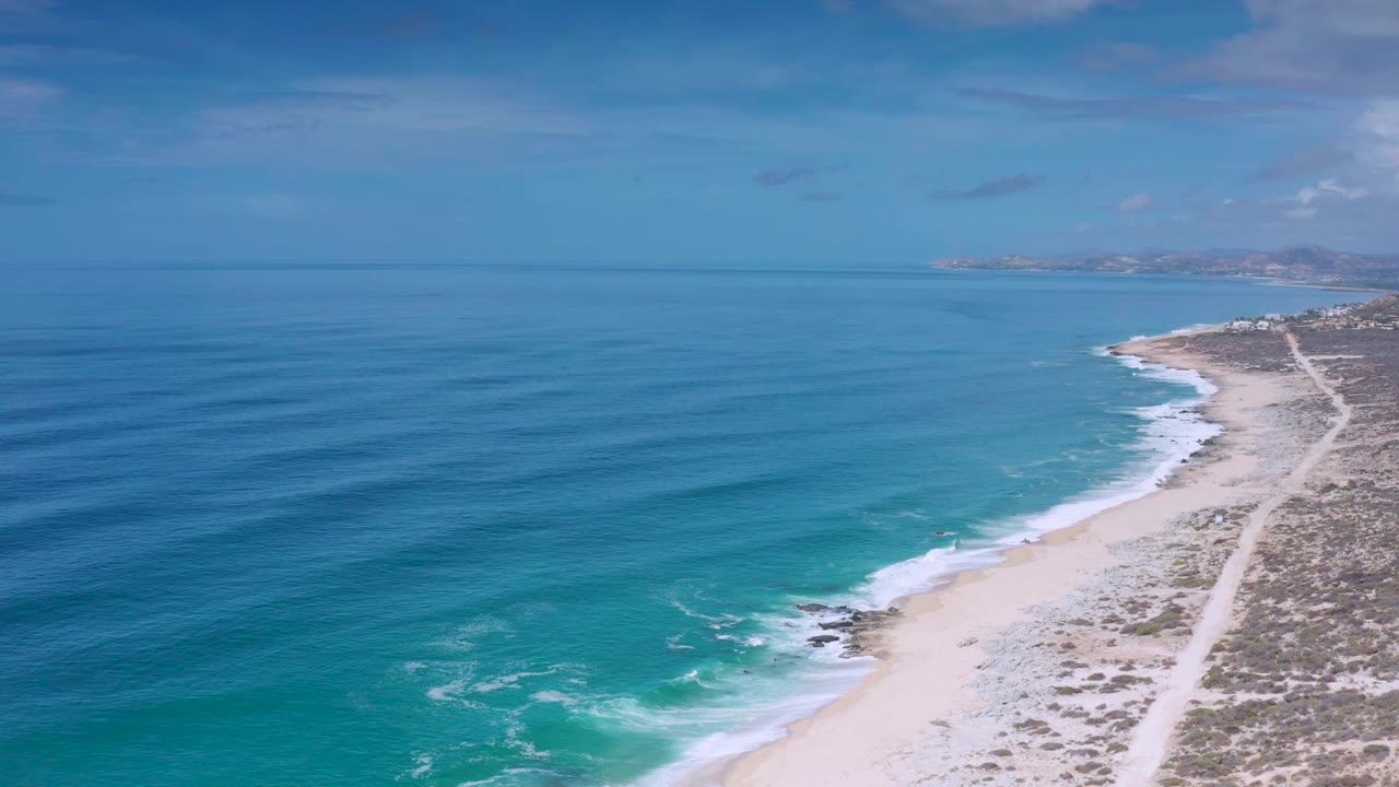 Elevated Los Cabos shoreline view with turquoise shallows near the beach, darker blue water offshore, scrubland to the right, and no visible people.