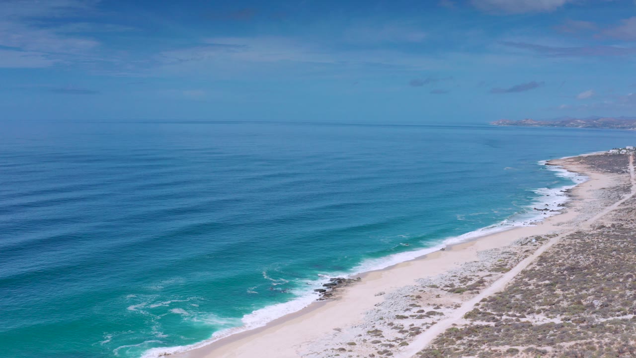 High aerial view over the Los Cabos coast, with deep blue ocean on the left, white surf along a pale beach, scrubby land to the right, and no visible people.