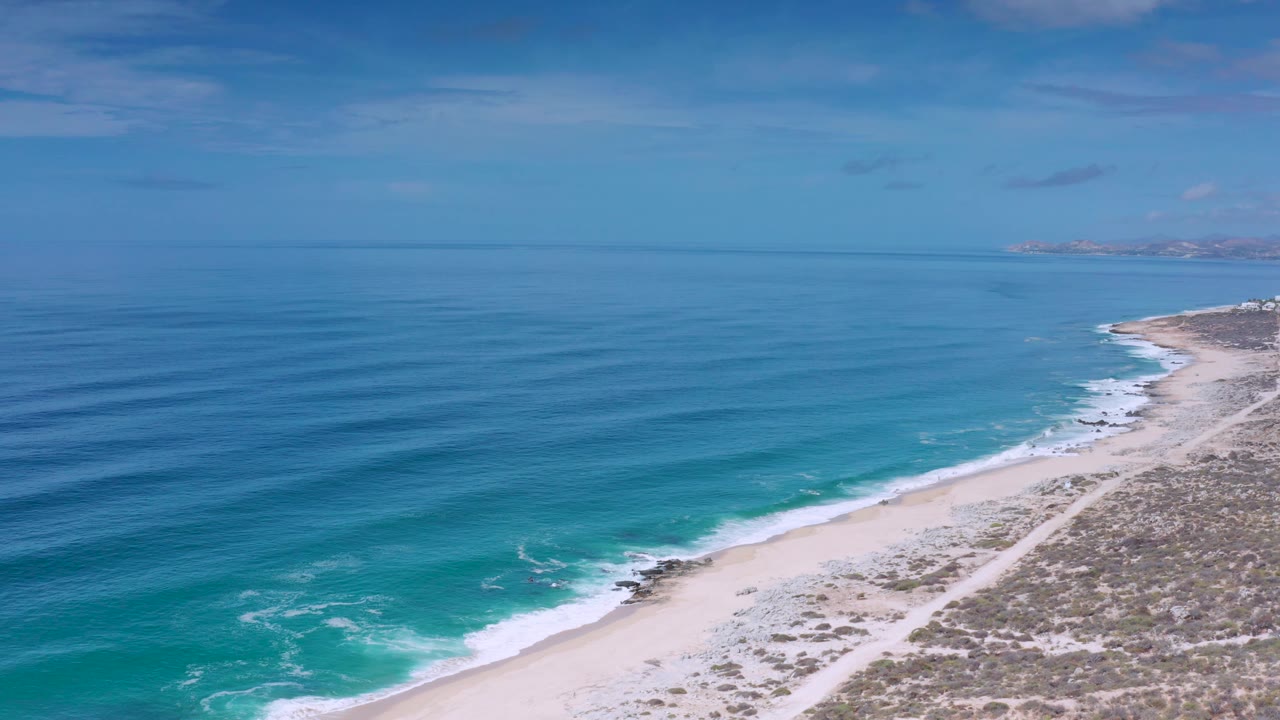 Oblique aerial frame of Los Cabos coastline, with long ocean swells moving toward a narrow beach, dry scrubland at frame right, distant land at the horizon, and no visible people.