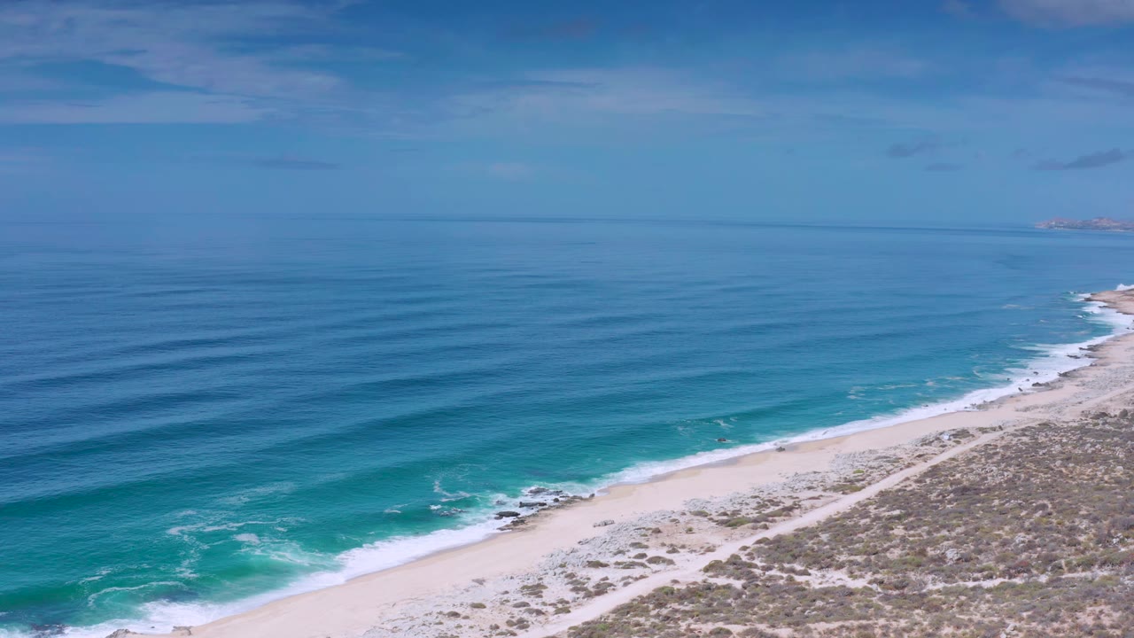 Los Cabos, Mexico beach seen from above as long blue wave bands approach the sand, with dune scrub in the lower right and no visible people.