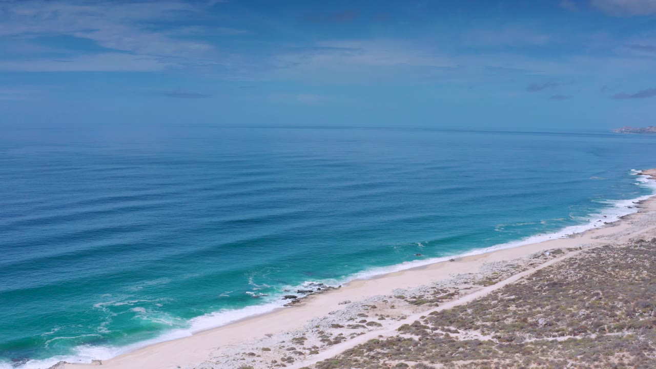 A wide Los Cabos, Mexico coastline frame shows surf tracing the beach edge, open water across most of the image, low clouds, and no visible people.