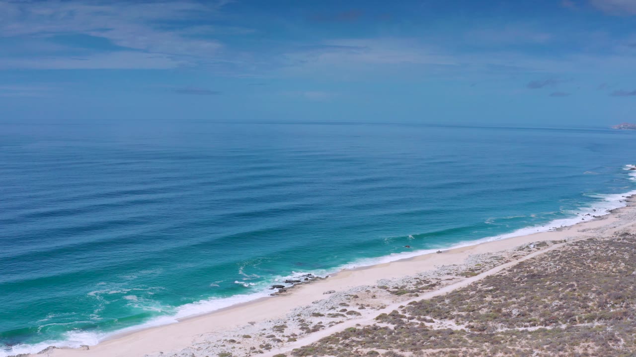 Mid-clip frame over the Los Cabos, Mexico shore with blue ocean on the left, white surf along the sand, scrub dunes to the right, and no visible people.