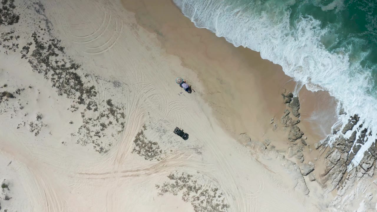 Overhead Los Cabos beach view with the foam line curling farther across the upper right, tire tracks arcing through the sand, rocks near the water, and no visible people.