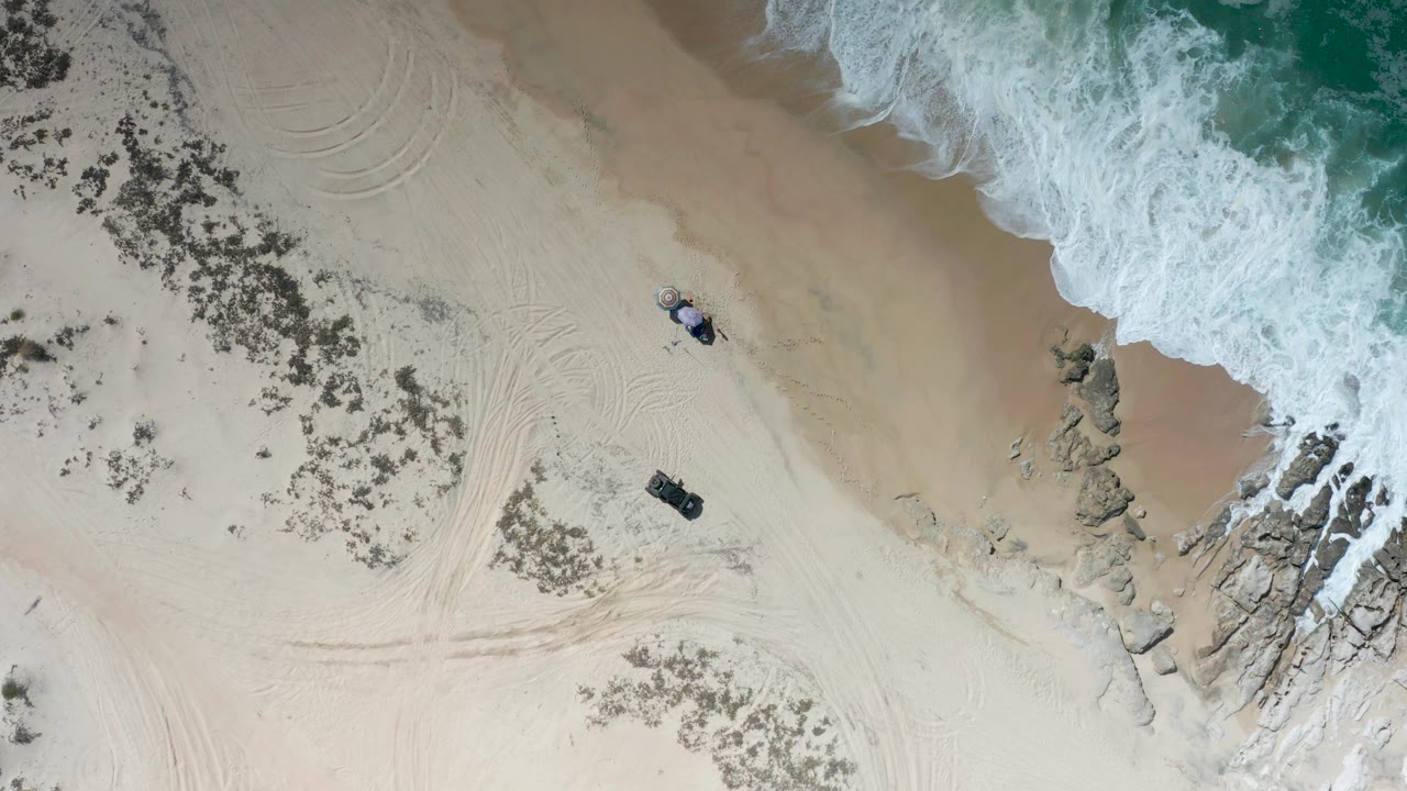 Overhead drone view of the Los Cabos shoreline with a wider band of wet sand near the water, white surf at the upper right, rocks on the right edge, and no visible people.