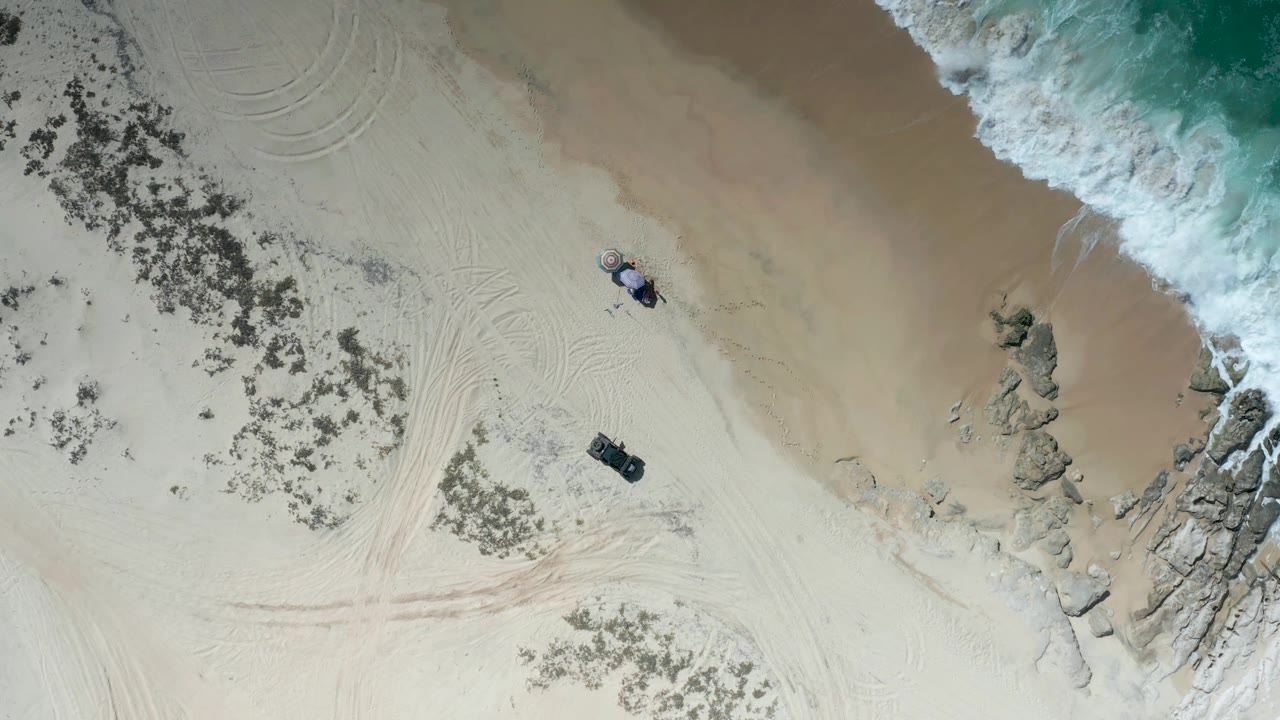 Aerial beach frame with waves approaching rocks and vehicles on sand.