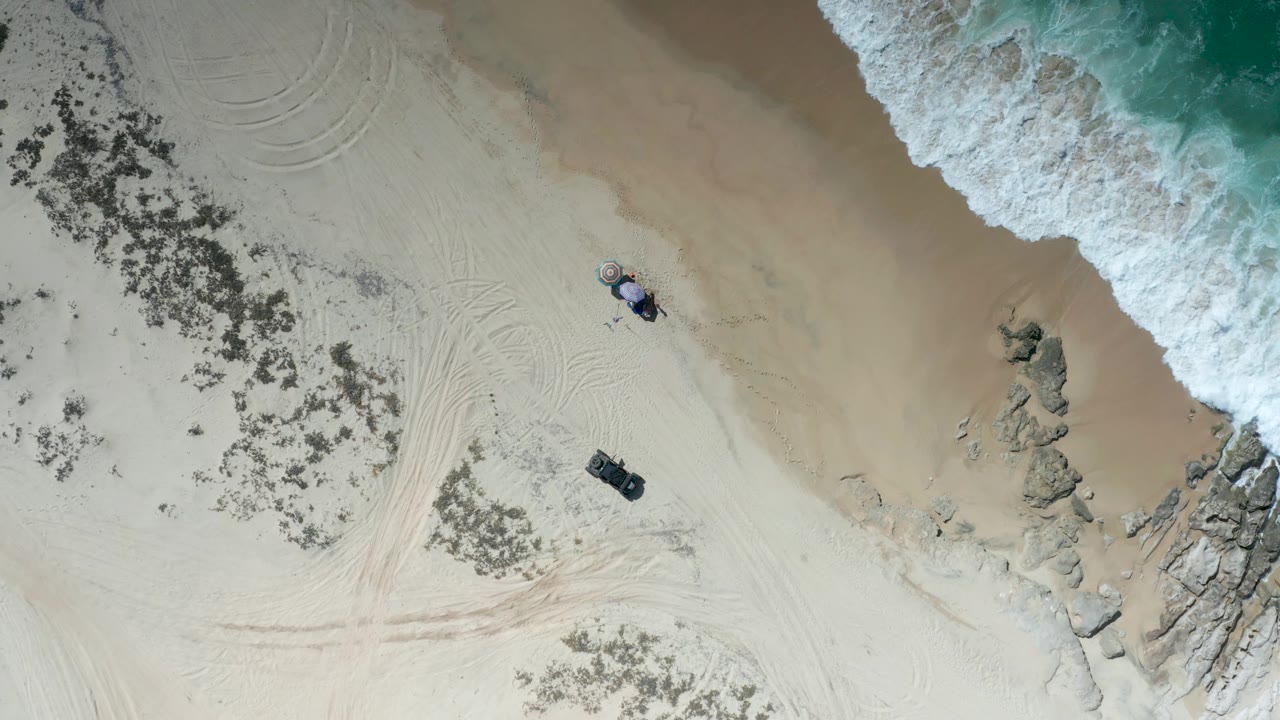 Top-down Los Cabos shoreline view with a larger band of foam at the upper right, rocks along the waterline, two parked vehicles on dry sand, and small people visible near the beach access point.