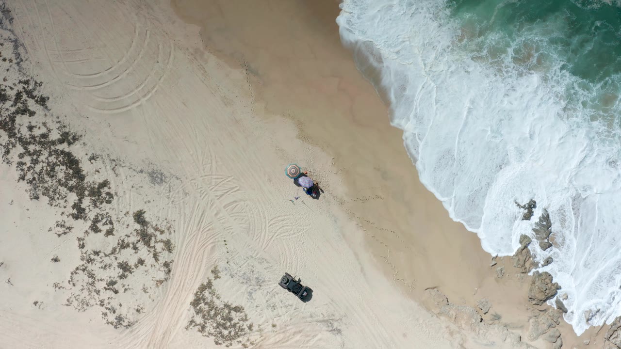 Top-down Los Cabos shoreline frame with foamy water along the upper right, broad pale sand through the center, curved track marks on the left, small beach gear below center, and no visible people.