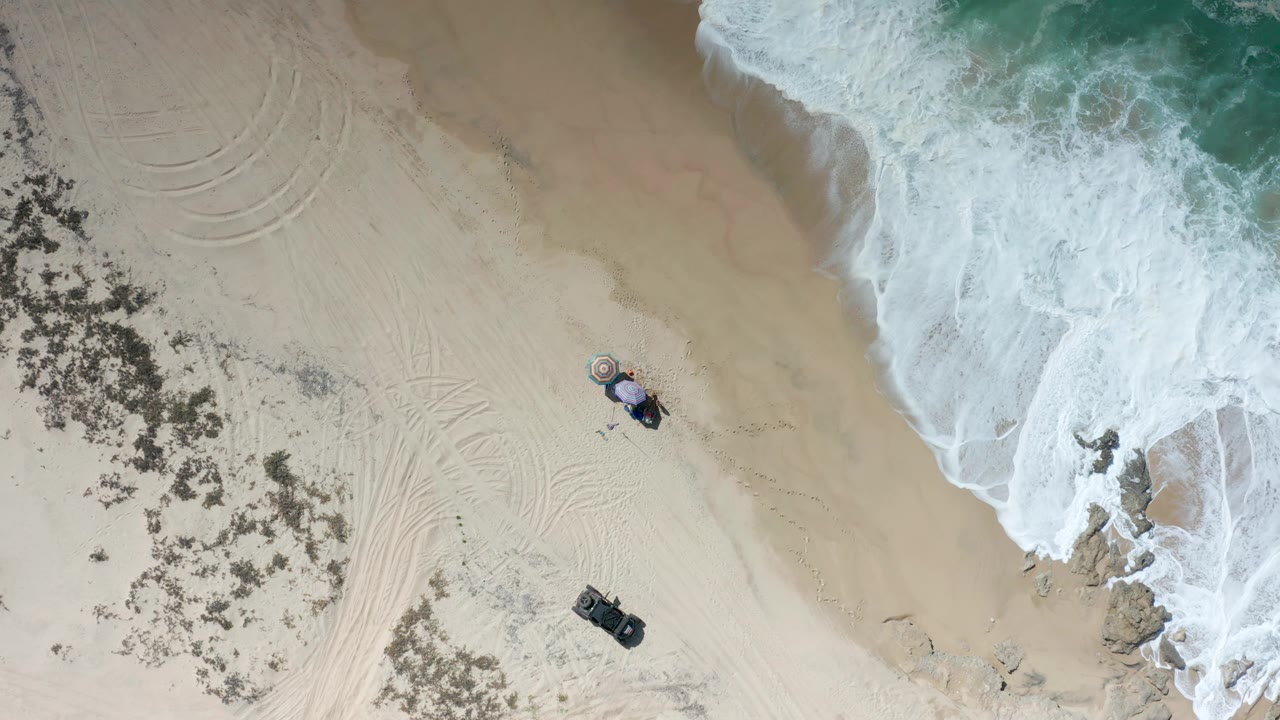 Aerial Los Cabos beach view with the surf shifted farther across the upper right, rocks partly surrounded by foam, open sand in the middle, a small setup near the lower center, and no visible people.