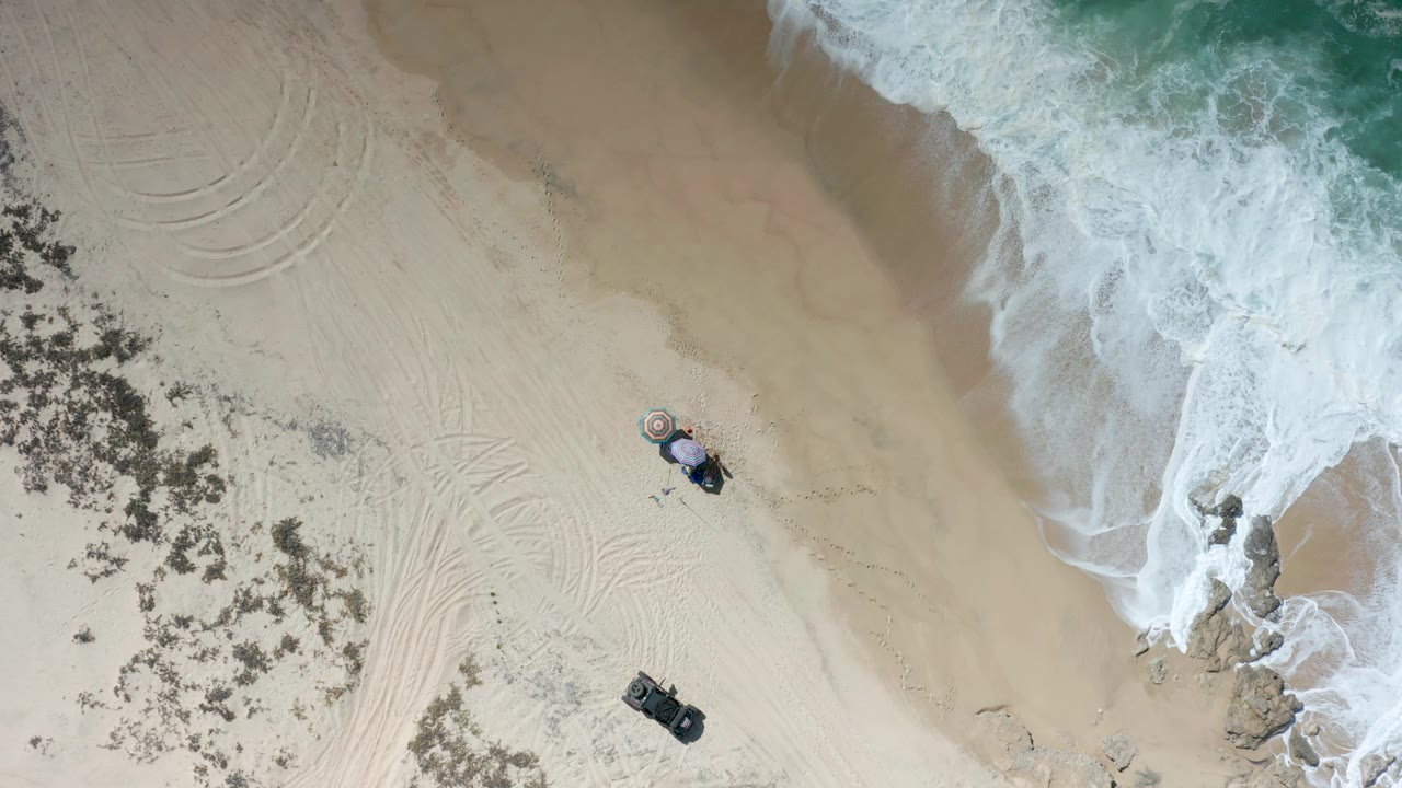 Overhead Los Cabos frame showing broad wet and dry sand, whitewater filling the right edge, curved tracks on the left, small beach objects near the lower middle, and no visible people.