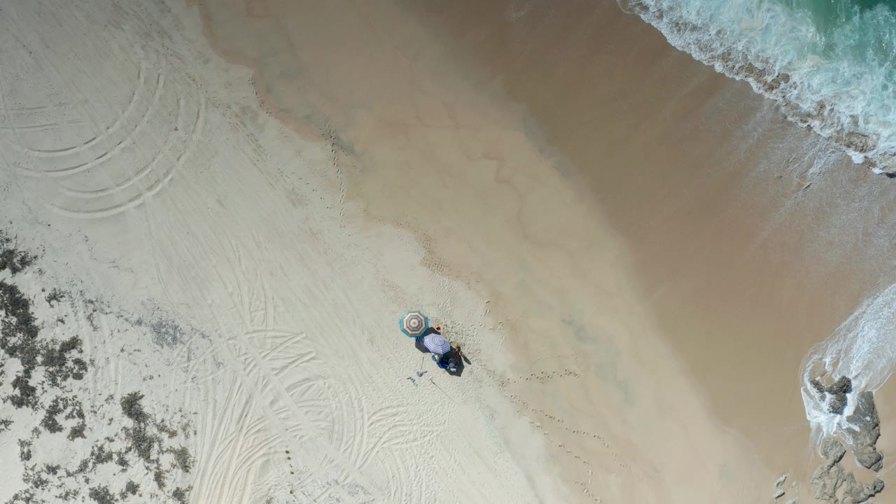 Overhead view of a Los Cabos beach with pale sand, surf foam along the upper edge, a rock at right, beach gear near the lower center, and no visible people.