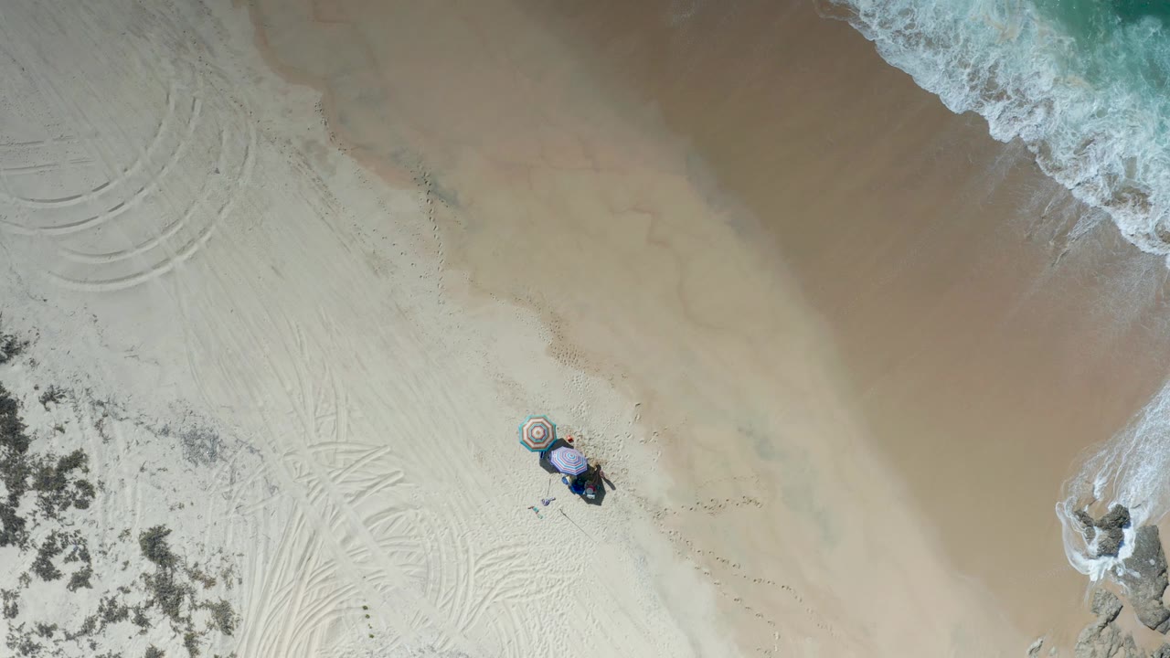 Overhead frame from Los Cabos with foamy water crossing the upper right, broad tan sand through the middle, an umbrella setup near the right side, and no visible people.