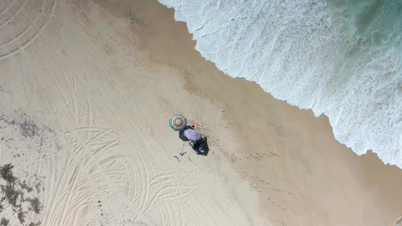 Overhead view in Los Cabos, Mexico, showing white surf at the top edge, pale sand with tire tracks, two striped umbrellas, beach gear, and small people near the shade.