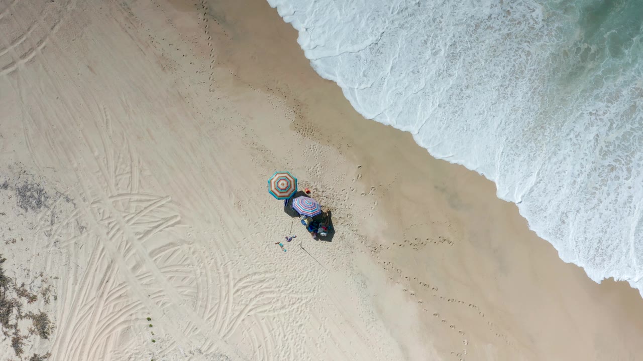 Aerial Los Cabos beach view with a band of white surf across the top, two colorful umbrellas near the lower center, beach gear, small people, and open sand marked by tracks.