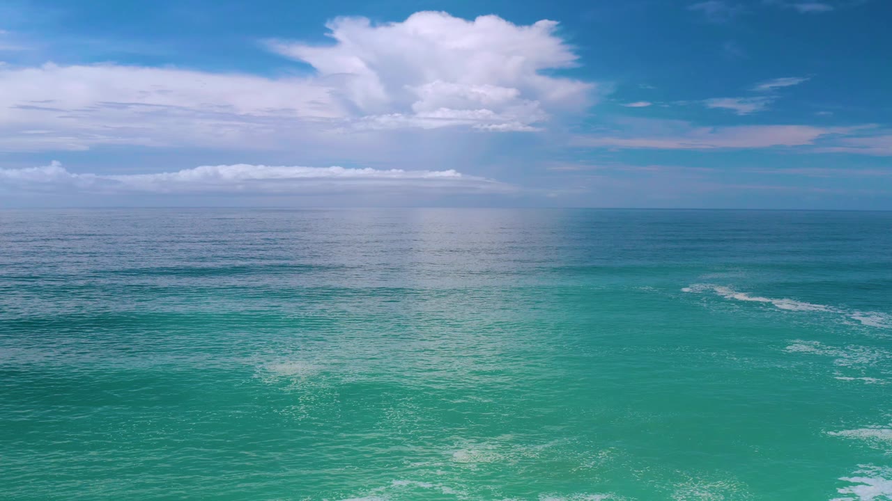 Los Cabos ocean horizon sits beneath layered white clouds, with turquoise water and faint wave breaks visible, no visible people.
