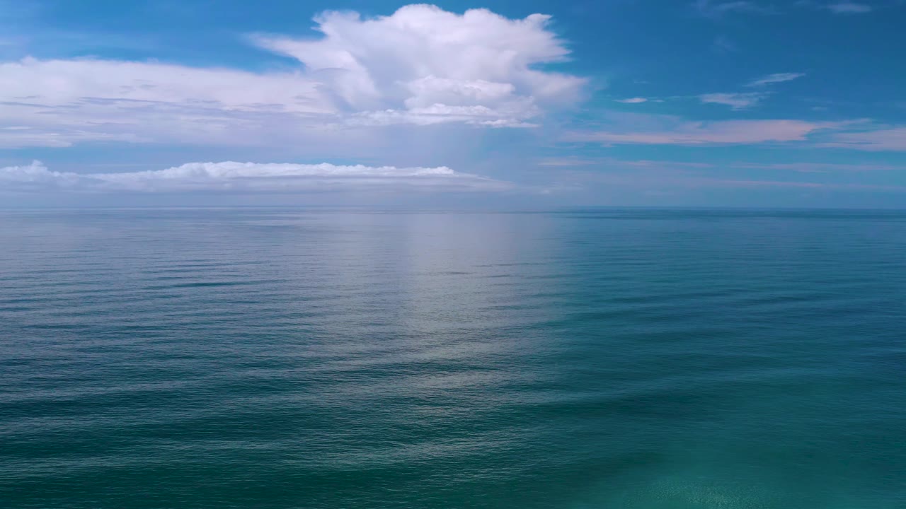 A wide Los Cabos, Mexico ocean frame with a low band of white clouds near the horizon, brighter cloud mass above, reflective water below, and no visible people.