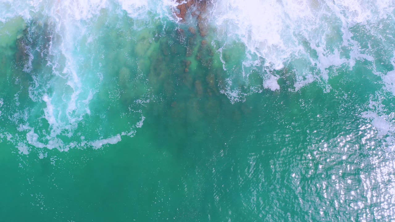 Overhead view in Los Cabos, Mexico of turquoise water and white foam spreading across reddish rocks near the top of frame, with no visible people.