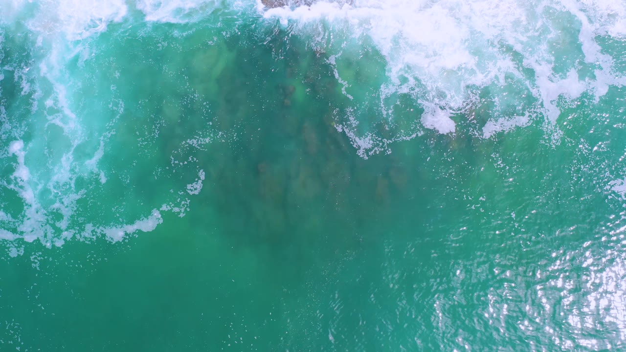 Overhead Los Cabos ocean view with green-blue water, a broken white surf line at the top, darker submerged areas, and no visible people.