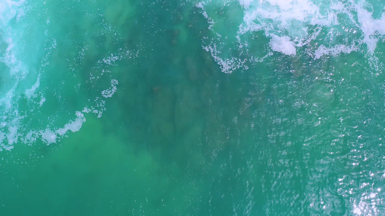 Clear green-blue sea in Los Cabos viewed from above, with a curved band of white foam, rippled sunlight, faint seabed color variation, and no visible people.