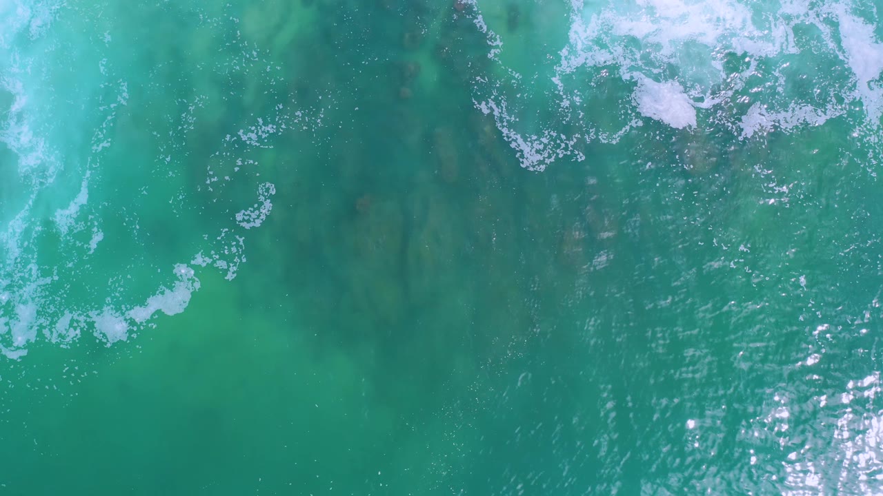 Top-down Los Cabos ocean surface with bright sunlight reflecting near the center, scattered white foam at the top, teal water, and no visible people.