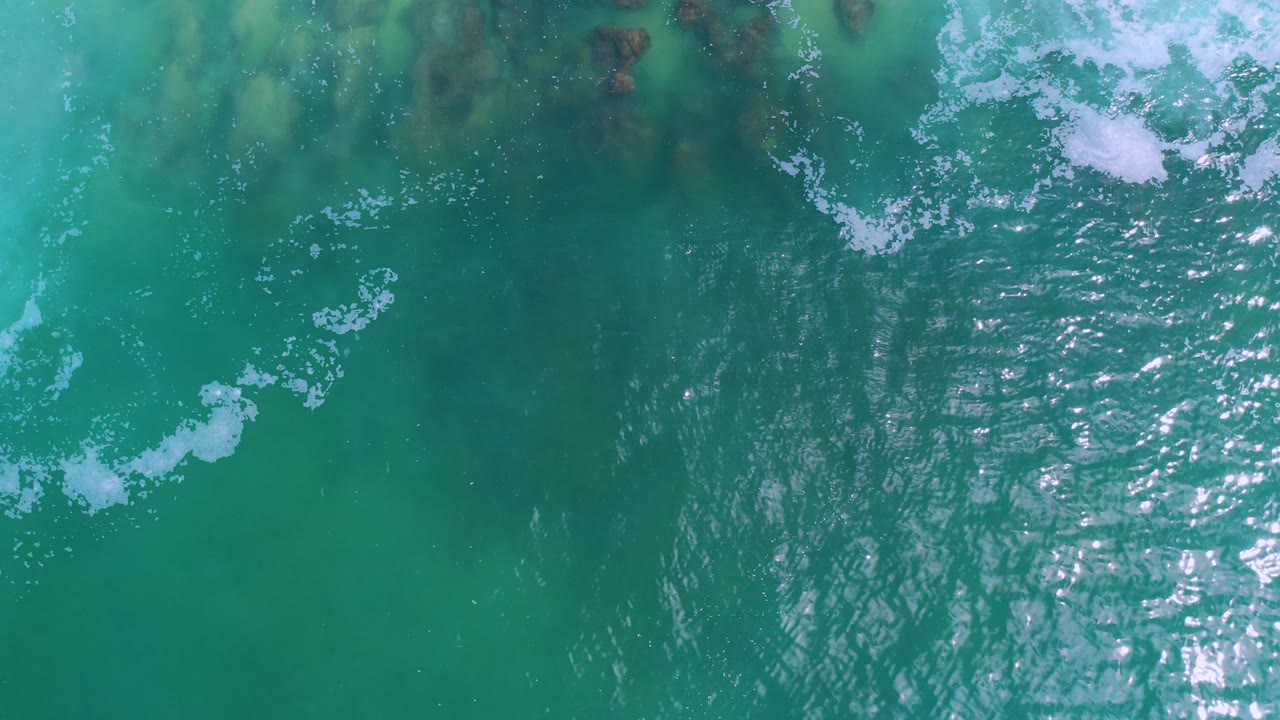Overhead view of turquoise ocean water in Los Cabos, Mexico, with white foam near the top edge, sun glare on ripples, darker underwater patches, and no visible people.