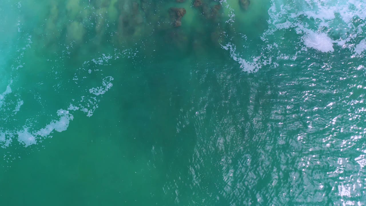 Aerial-style view of Los Cabos ocean water with scattered white foam on the upper right, a vertical band of sunlit ripples, muted green-blue water, and no visible people.