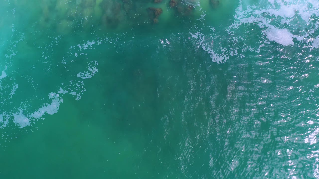 Overhead Los Cabos ocean frame with bright specular highlights down the right side, small white foam clusters near the top, green-blue water, and no visible people.