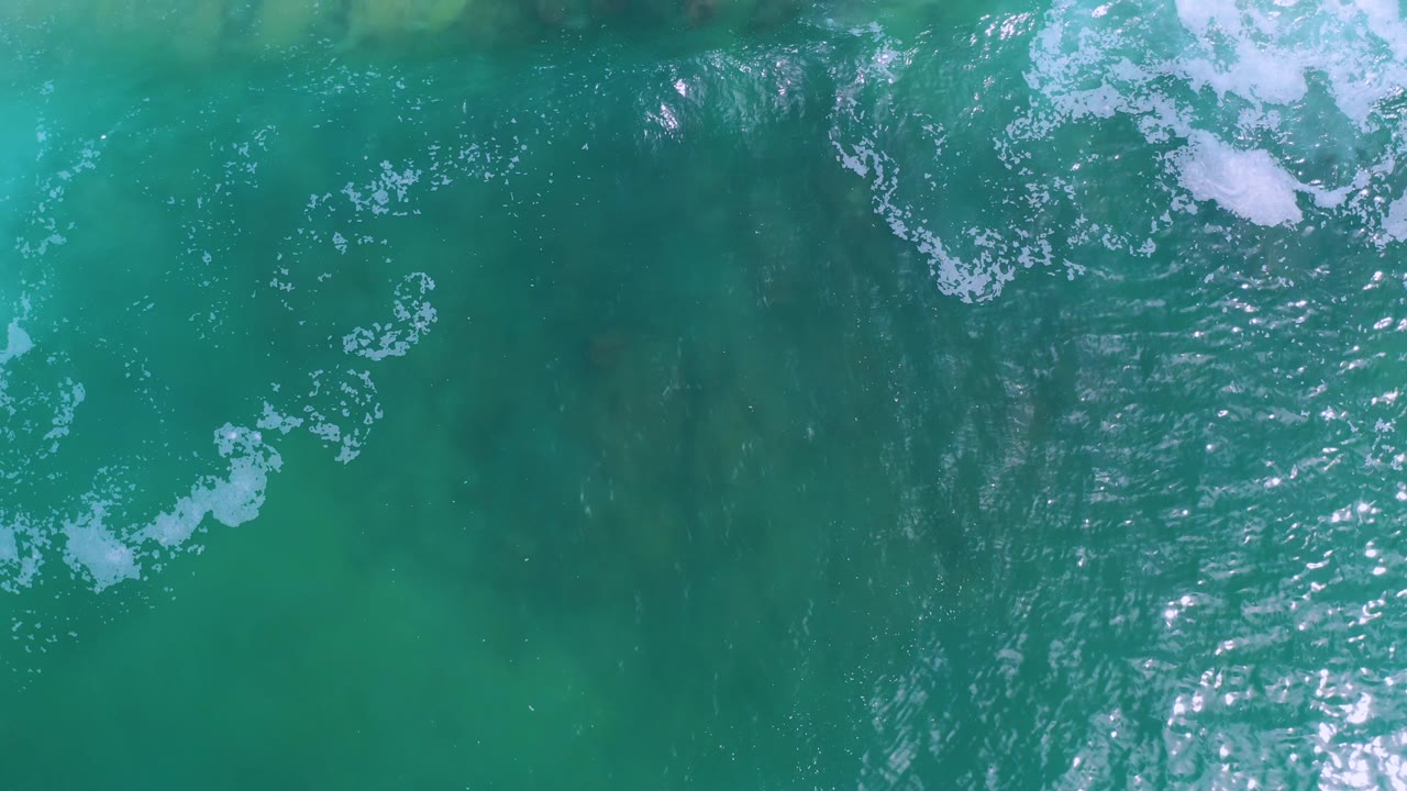 A top-down view of turquoise water in Los Cabos, Mexico, where white foam crosses the upper right and lower left areas, with soft underwater shadows and no visible people.