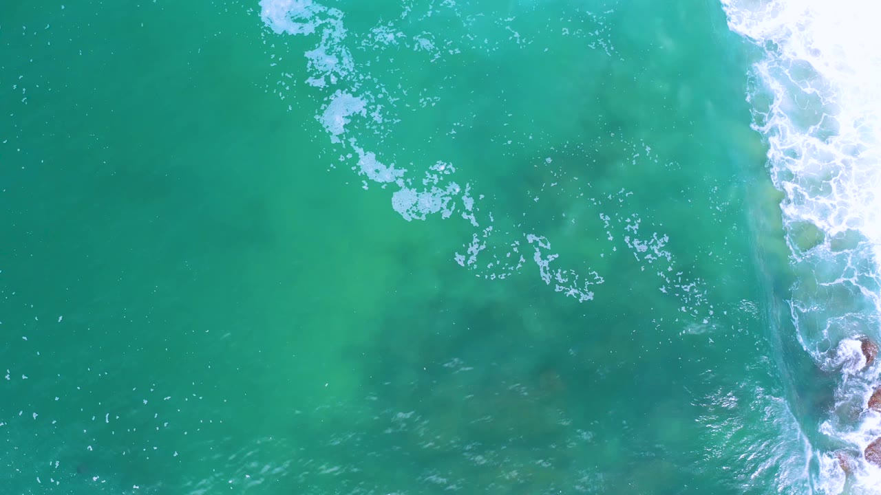 Overhead view of turquoise-green sea in Los Cabos, Mexico, with white surf breaking along the right edge and no visible people.