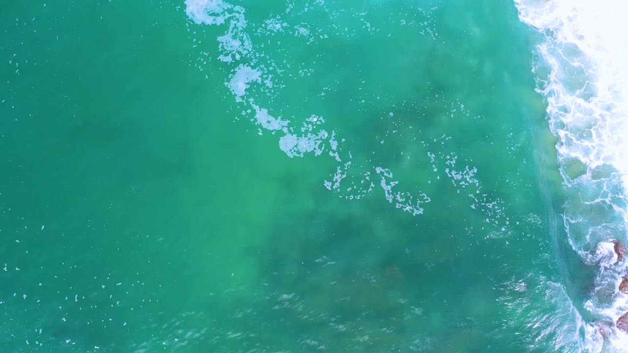 Aerial view over Los Cabos sea water with scattered foam clusters near the upper center and a narrow band of surf on the right, with no visible people.