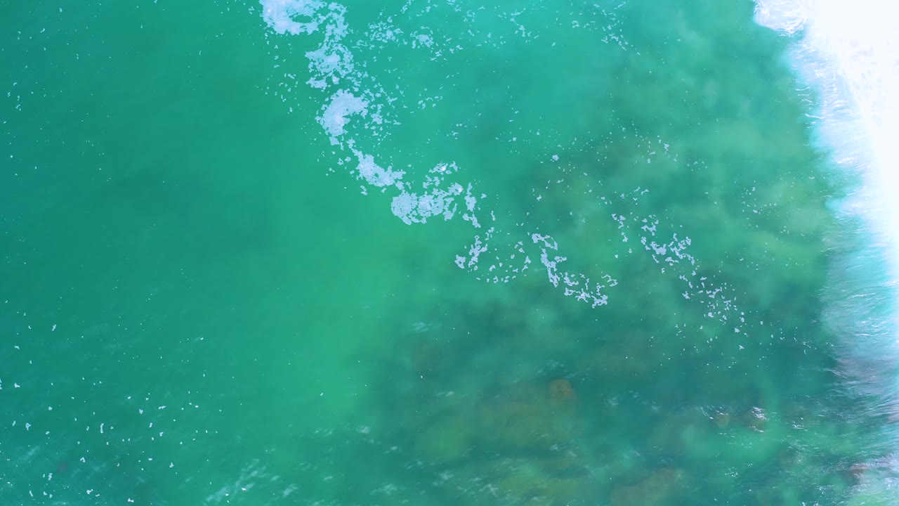 Overhead Los Cabos water frame with milky turquoise color, scattered white foam, faint submerged rock shapes near the lower right, and no visible people.