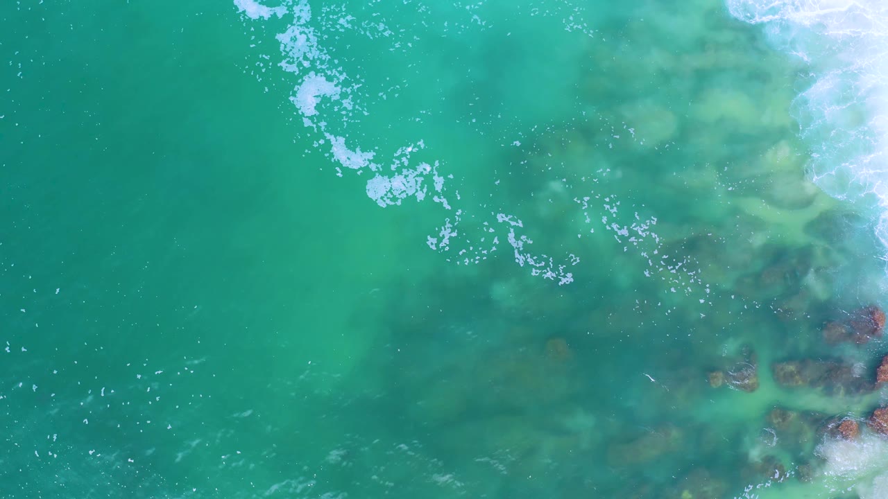 Aerial frame over Los Cabos water showing pale foam drifting across green-blue ocean and dark rock patches under the surface, with no visible people.