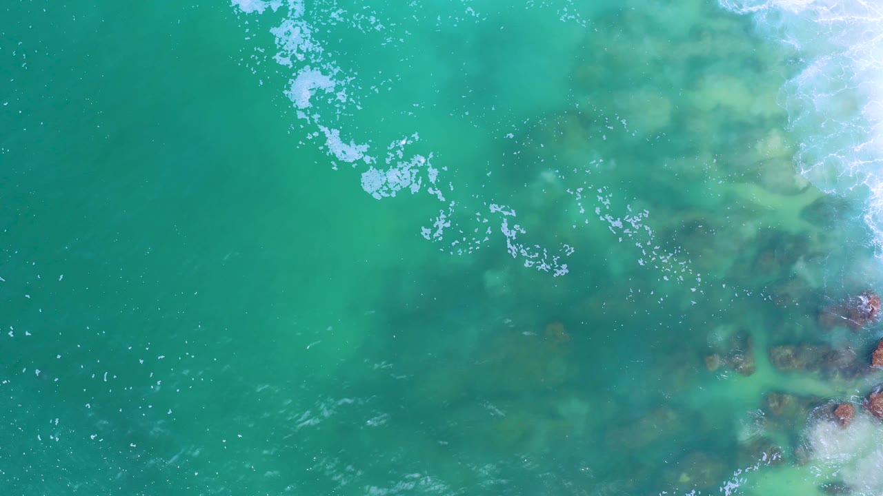 Top-down Los Cabos shoreline view with open turquoise water on the left, white foam near the top, and reddish-brown rocks under breaking surf on the right, with no visible people.