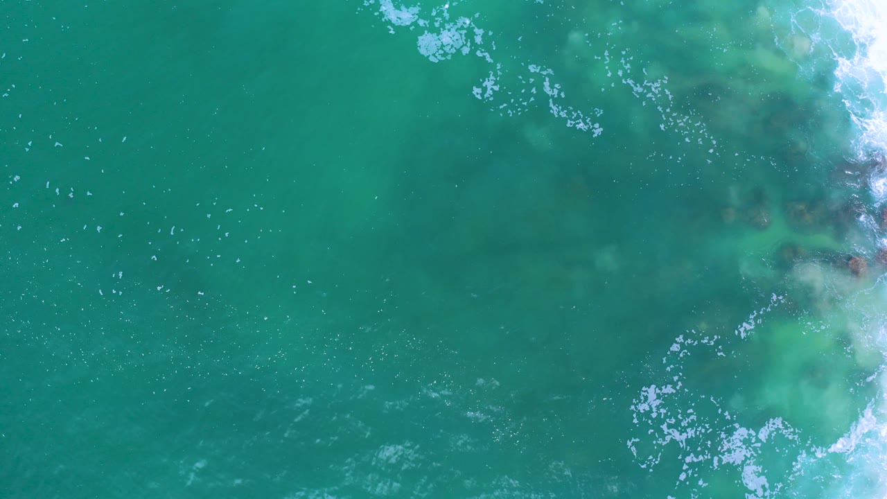 Overhead view of teal ocean water in Los Cabos, Mexico, with white foam entering along the right side and no visible people.